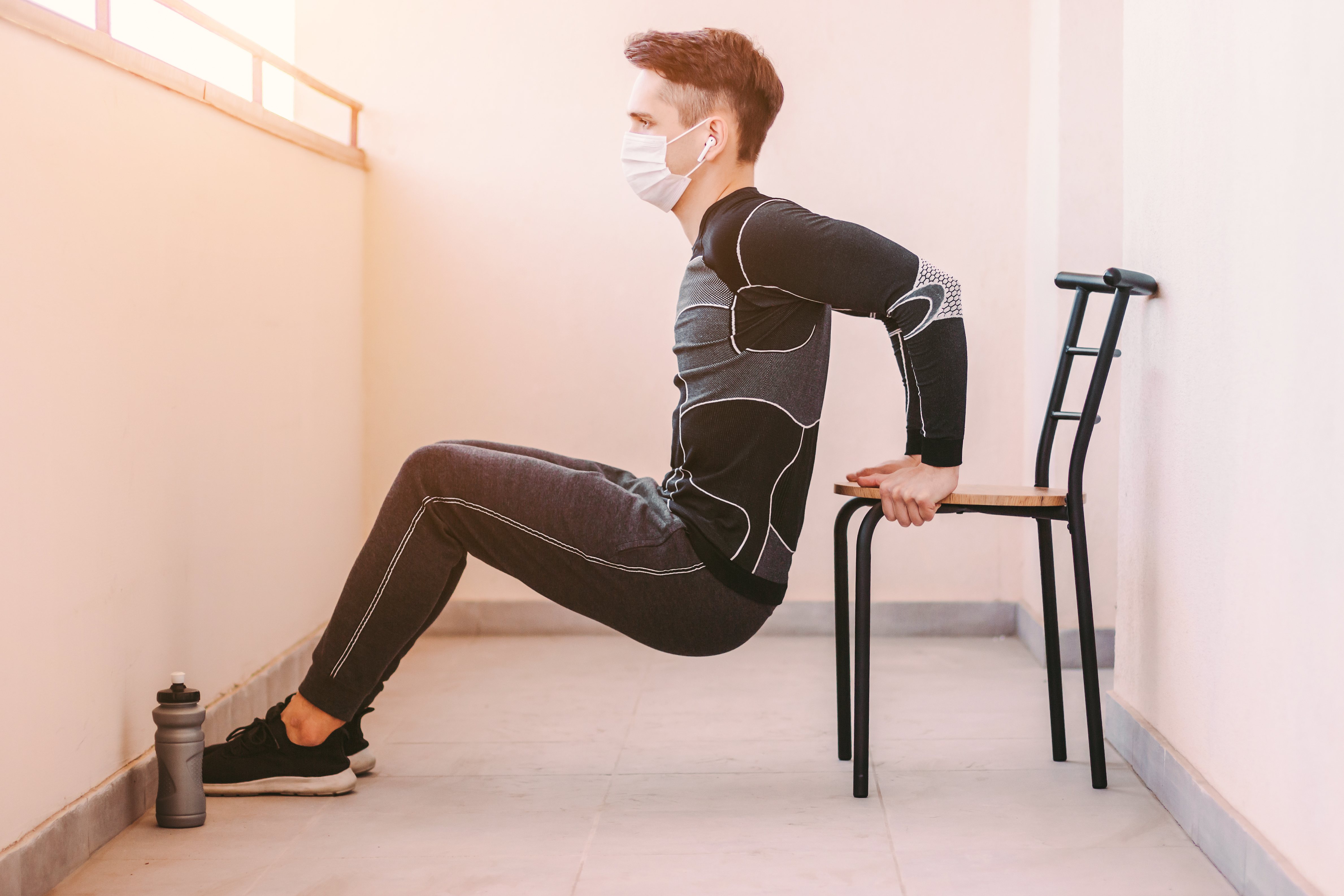 Joven deportista de confianza en la cara con mascarilla médica haciendo zambullidos triceps en la silla en casa.