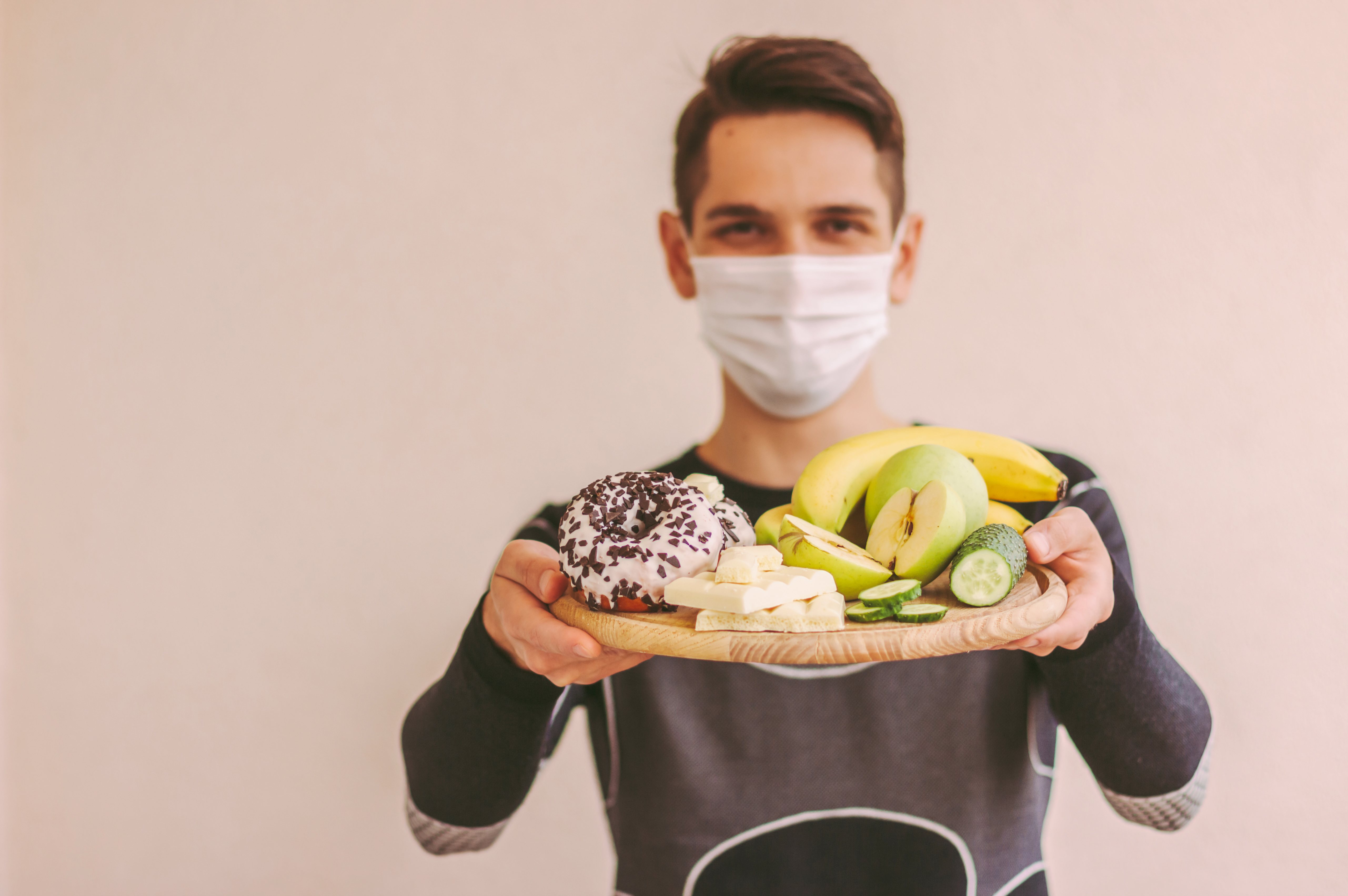 Feliz deportista con mascarilla médica sosteniendo tablón de madera con verduras, frutas frescas, donut y chocolate.